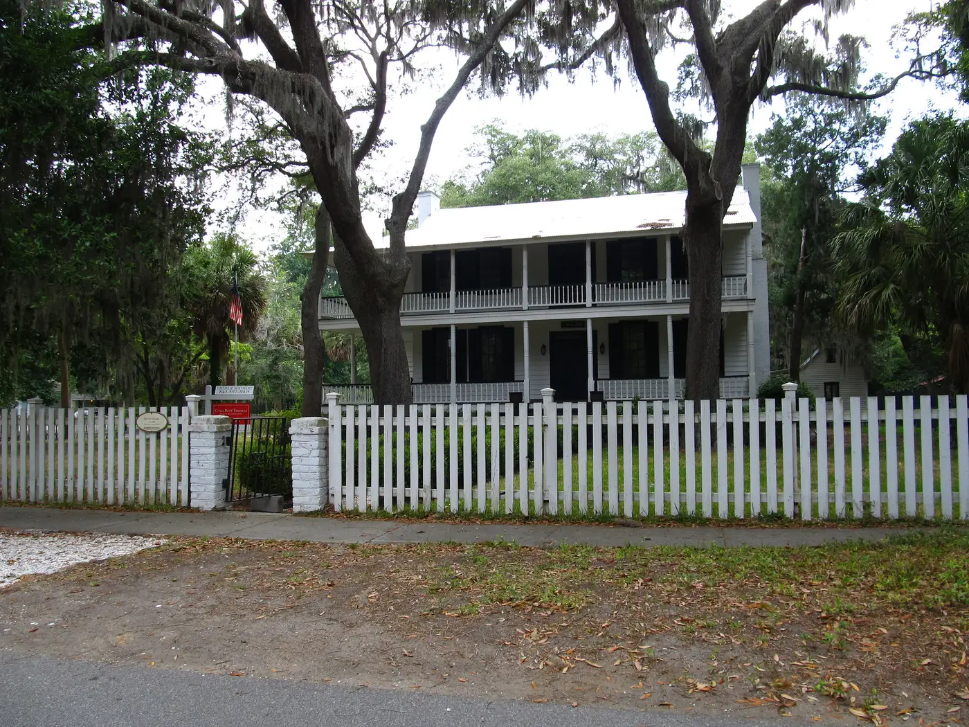 A two-story house behind a white picket fence under live oaks in Old Town Bluffton.